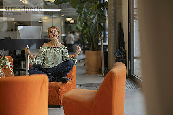 Lächelnde reife Geschäftsfrau sitzt mit gekreuzten Beinen auf einem orangefarbenen Stuhl im Büro