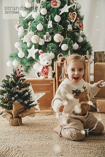 Glückliches Mädchen  das zu Hause in der Nähe von Weihnachtsbäumen spielt