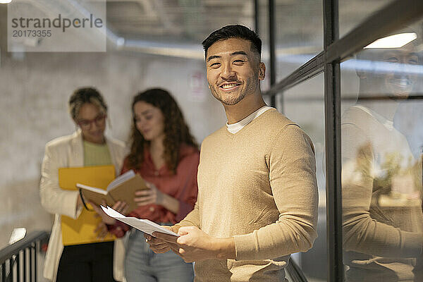 Lächelnder junger Geschäftsmann steht mit Dokument neben Kollegen und diskutiert im beleuchteten Flur