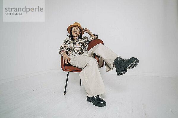 Sorglose ältere Frau sitzt auf einem Stuhl vor einer weißen Wand