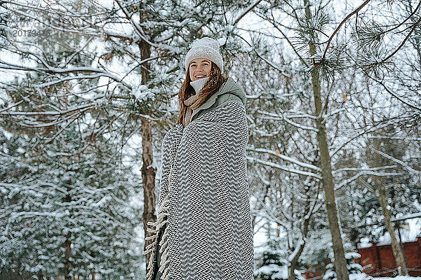 Lächelnde Frau  in eine Decke gehüllt  steht im Winter neben Bäumen