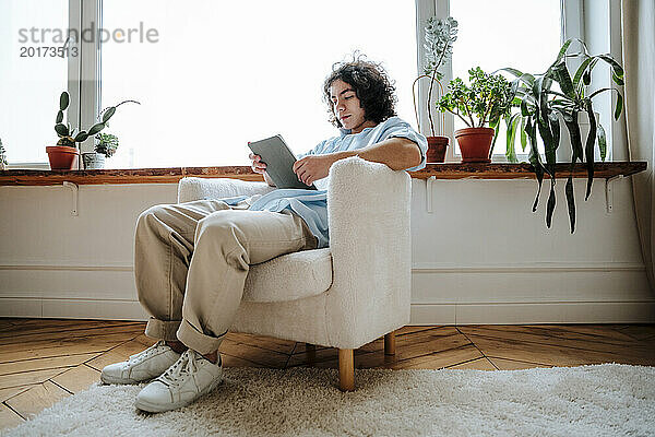 Mann sitzt auf einem Sessel und benutzt zu Hause einen Tablet-PC