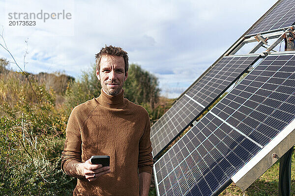 Lächelnder Mann steht an einem sonnigen Tag mit Smartphone in der Nähe eines Solarpanels