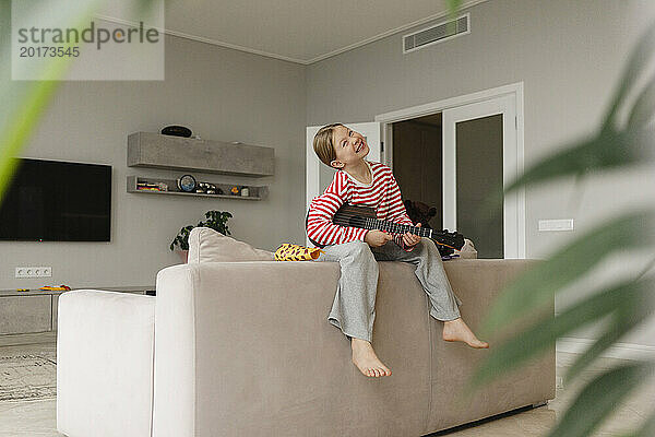 Glückliches Mädchen  das auf dem Sofa sitzt und zu Hause Spielzeuggitarre spielt