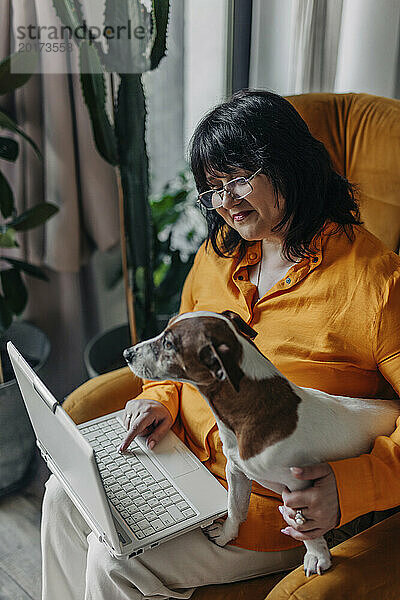 Lächelnde reife Frau sitzt mit Hund und Laptop auf einem Sessel zu Hause