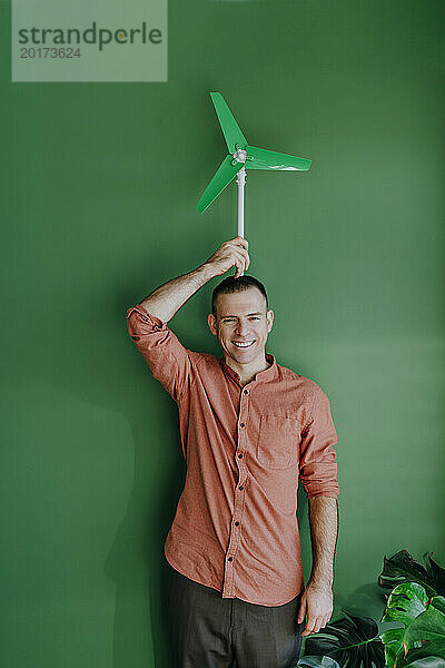 Glücklicher Freiberufler hält Windturbinenmodell auf dem Kopf vor grüner Wand