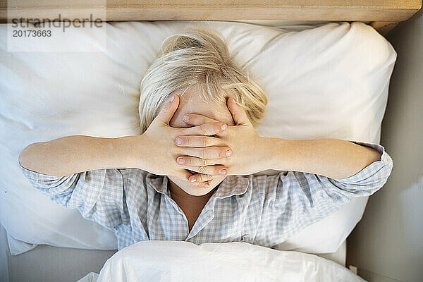 Junge bedeckt Gesicht mit Händen und liegt zu Hause auf dem Bett