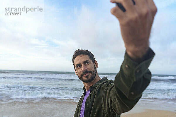 Mann macht Selfie mit Smartphone vor dem Meer am Strand