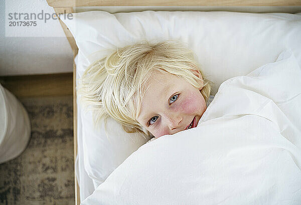 Lächelnder blonder Junge  der zu Hause auf dem Bett liegt