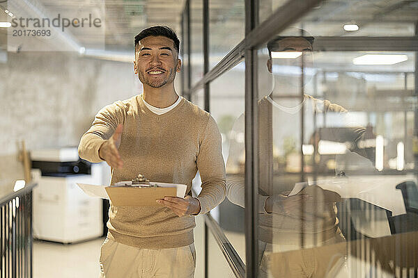 Lächelnder junger Geschäftsmann mit Dokumenten  der an der Glaswand im Büro zum Händedruck greift
