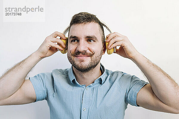 Lächelnder Mann hält kabellose Kopfhörer in der Hand und hört Musik vor der Wand