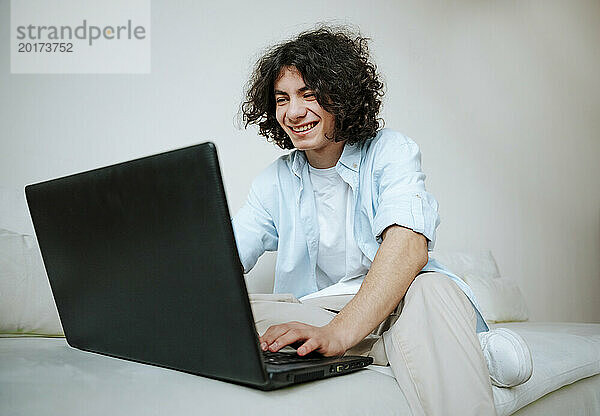 Glücklicher junger Mann mit lockigem Haar  der zu Hause auf dem Sofa einen Laptop benutzt