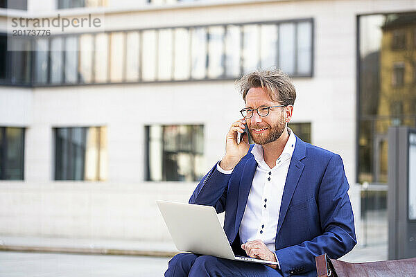 Lächelnder Geschäftsmann mit Laptop  der in der Stadt auf dem Smartphone spricht