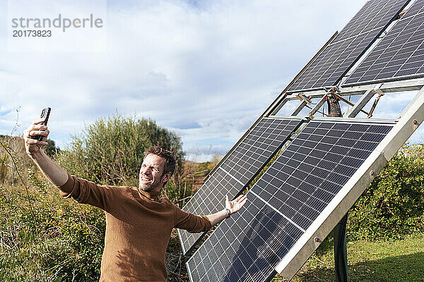 Glücklicher Mann  der an einem sonnigen Tag ein Selfie mit seinem Smartphone in der Nähe von Sonnenkollektoren zeigt und macht