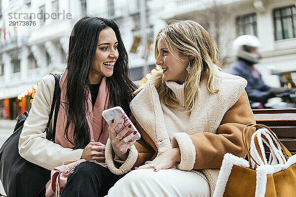 Lächelnde Frau hält Smartphone in der Hand und spricht mit einer Freundin  die auf einer Bank in der Stadt sitzt