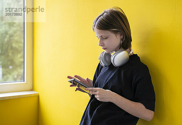 Mädchen mit kabellosen Kopfhörern benutzt Smartphone in der Nähe der gelben Wand zu Hause