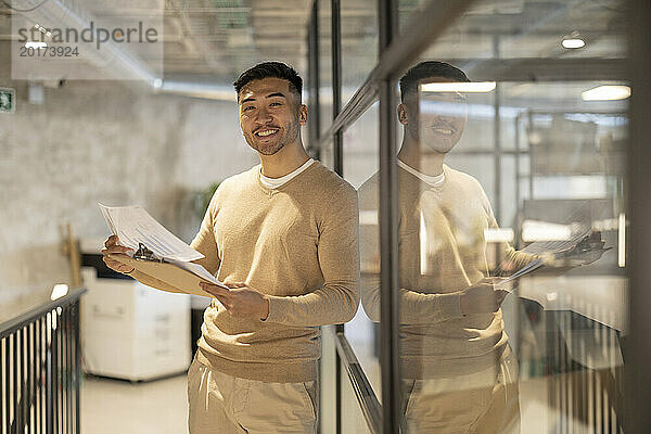 Lächelnder junger Geschäftsmann mit Dokumenten  der im Büro an einer Glaswand lehnt