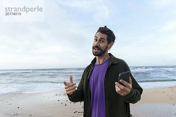 Glücklicher Mann gestikuliert und hält Smartphone am Strand