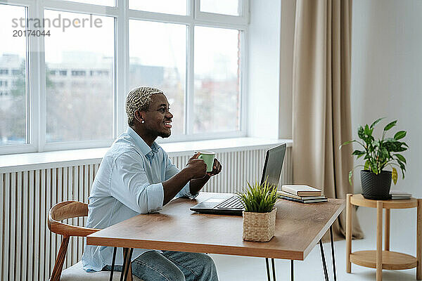 Lächelnder Freiberufler sitzt mit Kaffeetasse und Laptop am Schreibtisch