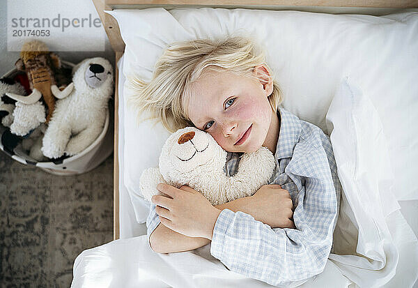 Lächelnder Junge liegt mit Teddybär zu Hause im Bett