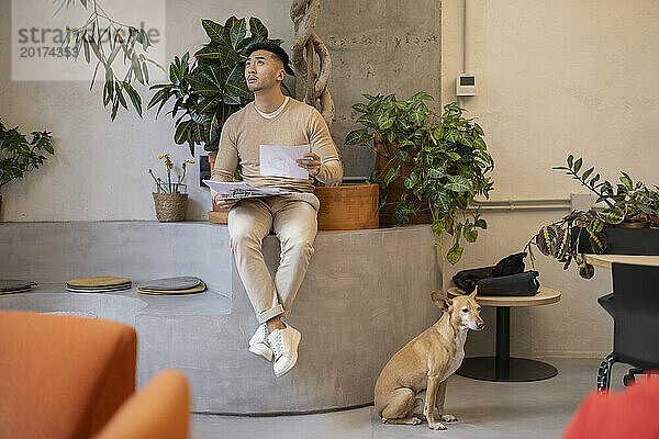 Junger Geschäftsmann sitzt mit Dokumenten neben Hund im Büro