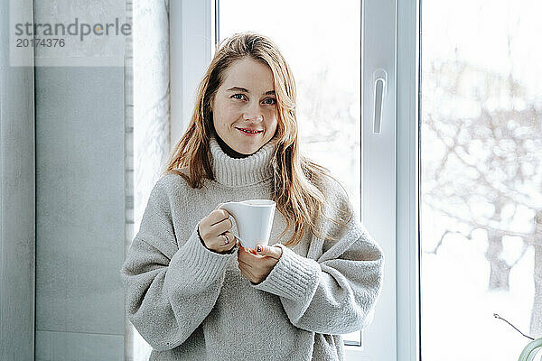 Lächelnde Frau mit braunen Haaren hält zu Hause eine Kaffeetasse am Fenster