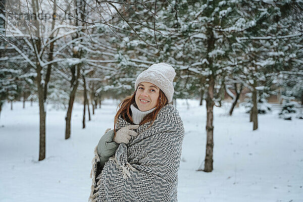 Lächelnde Frau in Decke gehüllt neben Bäumen im Schnee
