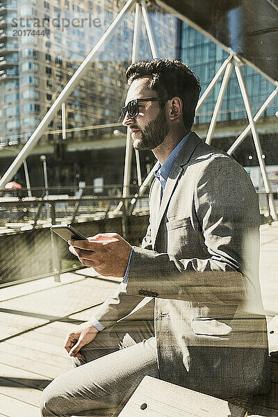 Geschäftsmann hält Smartphone in der Hand und sitzt an sonnigem Tag auf einer Bank in der Stadt