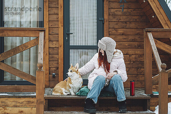 Glückliche Frau sitzt mit Corgi-Hund auf der Veranda einer Blockhütte
