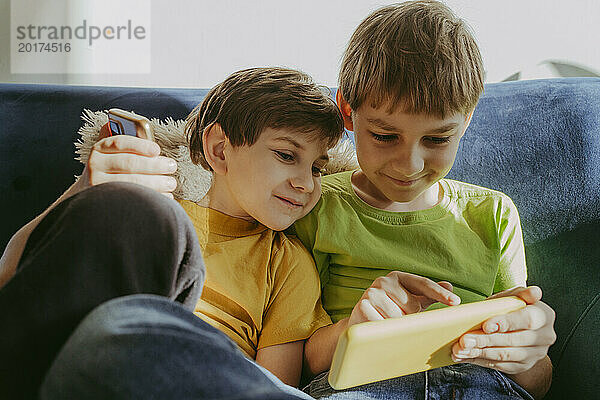 Lächelnde Brüder  die zu Hause auf dem Sofa sitzen und mit dem Smartphone spielen