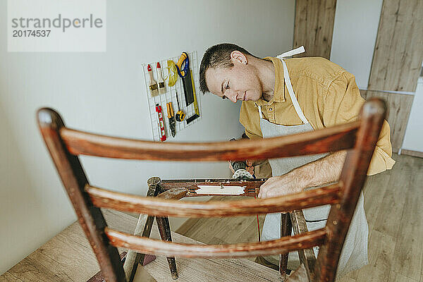 Man repairing old wooden chair using sander tool at home