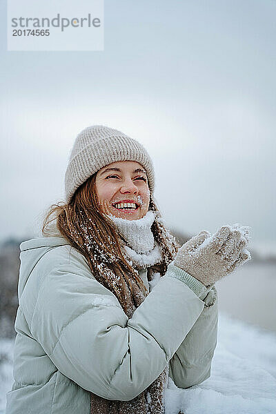 Glückliche Frau trägt warme Kleidung und spielt im Winter mit Schnee