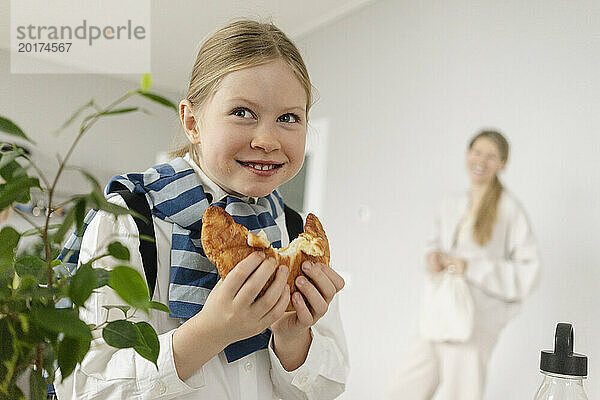 Verspieltes Mädchen isst Croissant mit Mutter im Hintergrund zu Hause