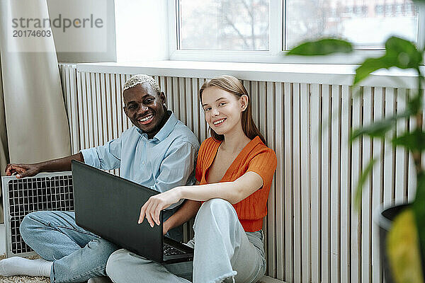 Glückliche Männer und Frauen sitzen mit Laptop am Fenster zu Hause