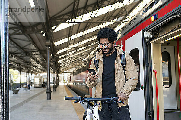 Lächelnder Mann benutzt Mobiltelefon und steht neben dem Fahrrad am Bahnhof in der Nähe des Zuges