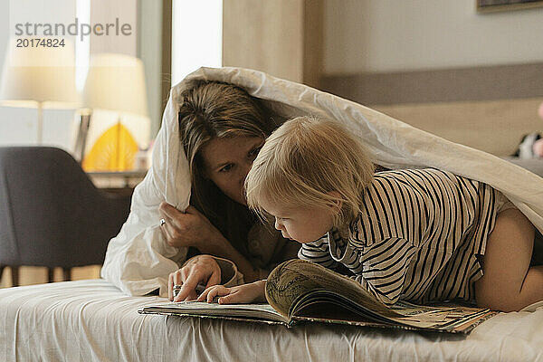 Mother reading book to daughter under blanket in bed at home