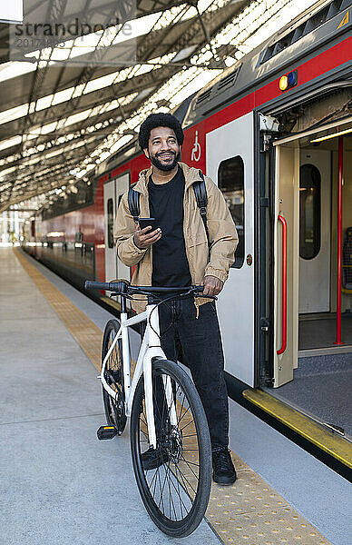 Lächelnder Mann hält Smartphone mit dem Fahrrad in der Nähe des Zuges am Bahnhof