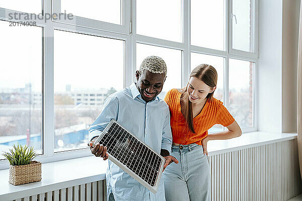 Glücklicher Mann hält Solarpanel in der Hand  während Frau zu Hause steht