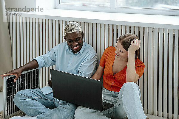 Lächelnde Freiberufler sitzen mit Laptop und Solarpanel am Fenster zu Hause