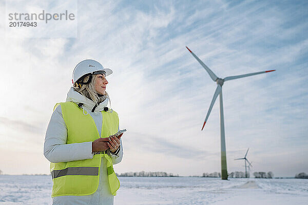 Ingenieur mit Schutzhelm und Smartphone steht im Windpark