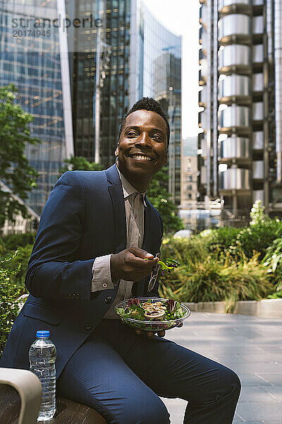 Lächelnder reifer Geschäftsmann sitzt mit einer Schüssel Salat im Büropark
