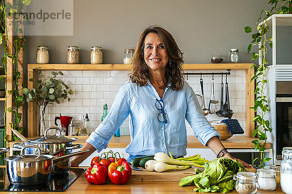 Lächelnde ältere Frau steht zu Hause neben Gemüse auf der Küchentheke