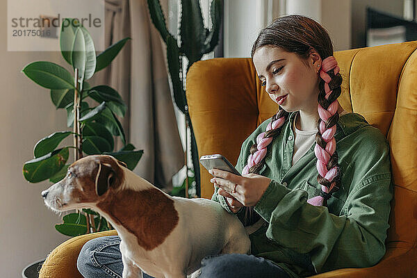 Lächelnde junge Frau sitzt mit Hund und benutzt zu Hause Smartphone