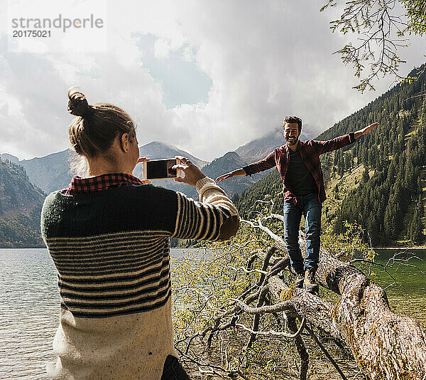 Junge Frau fotografiert einen glücklichen Mann  der auf einem umgestürzten Baumstamm in der Nähe des Vilsalpsees und der Berge in Tirol  Österreich  läuft