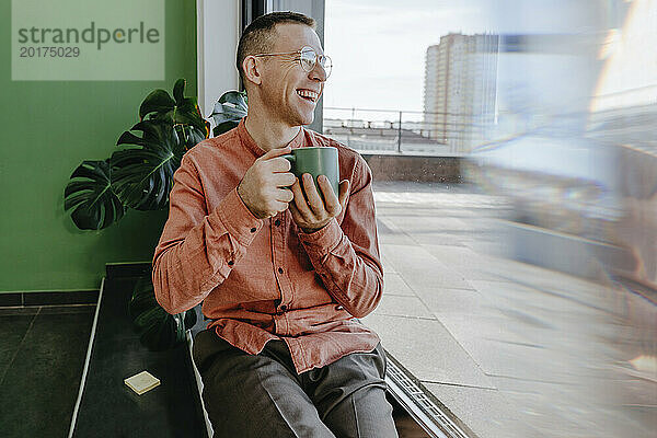 Glücklicher Mann sitzt mit Kaffeetasse in der Nähe der Glastür