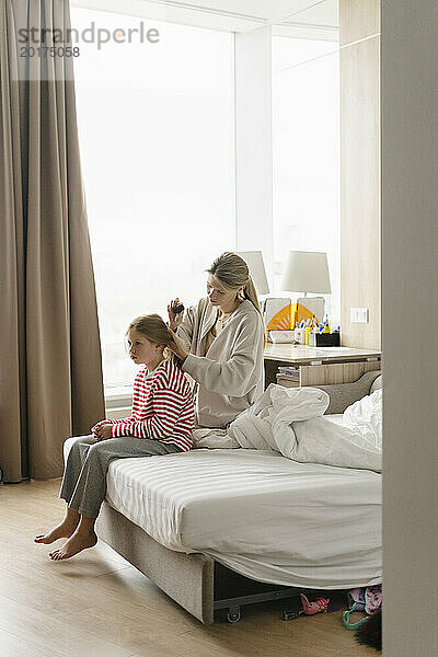 Mother tying daughter's hair sitting on bed at home