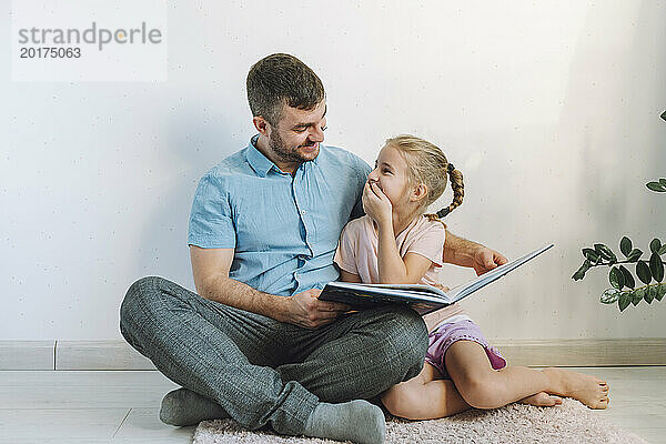 Glücklicher Vater liest seiner Tochter zu Hause vor der Wand ein Buch vor