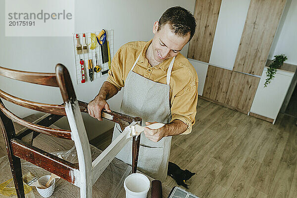 Smiling man painting chair with brush standing at home