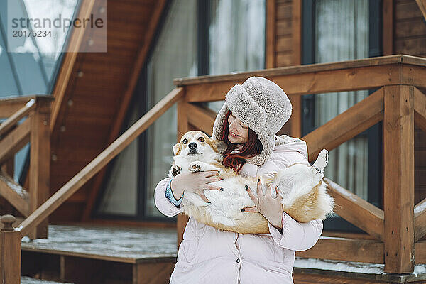 Lächelnde junge Frau hält Corgi-Hund vor Blockhütte