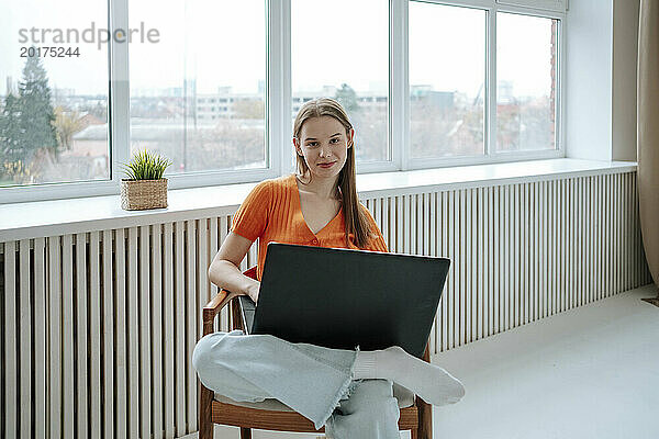 Lächelnder junger Freiberufler sitzt mit Laptop auf dem Sessel zu Hause